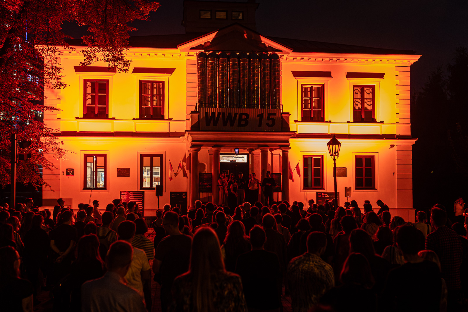 Zdjęcie Muzeum Woli wieczorem podczas wernisażu. Fasada oświetlona na pomarańczowo, przed budynkiem tłum ludzi.